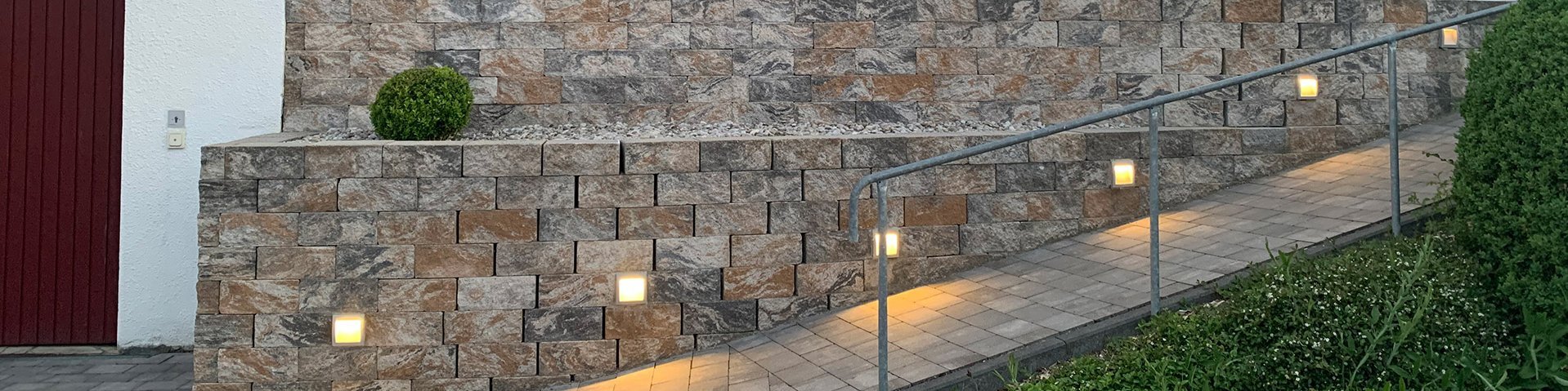 Ein Haus von der Seite. Davor steht eine Steinmauer mit Lichtern darin