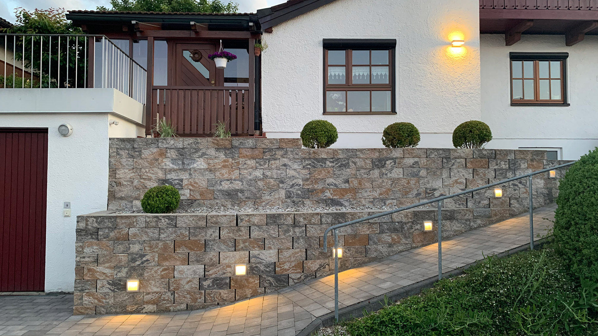 Ein Haus von der Seite. Davor steht eine Steinmauer mit Lichtern darin